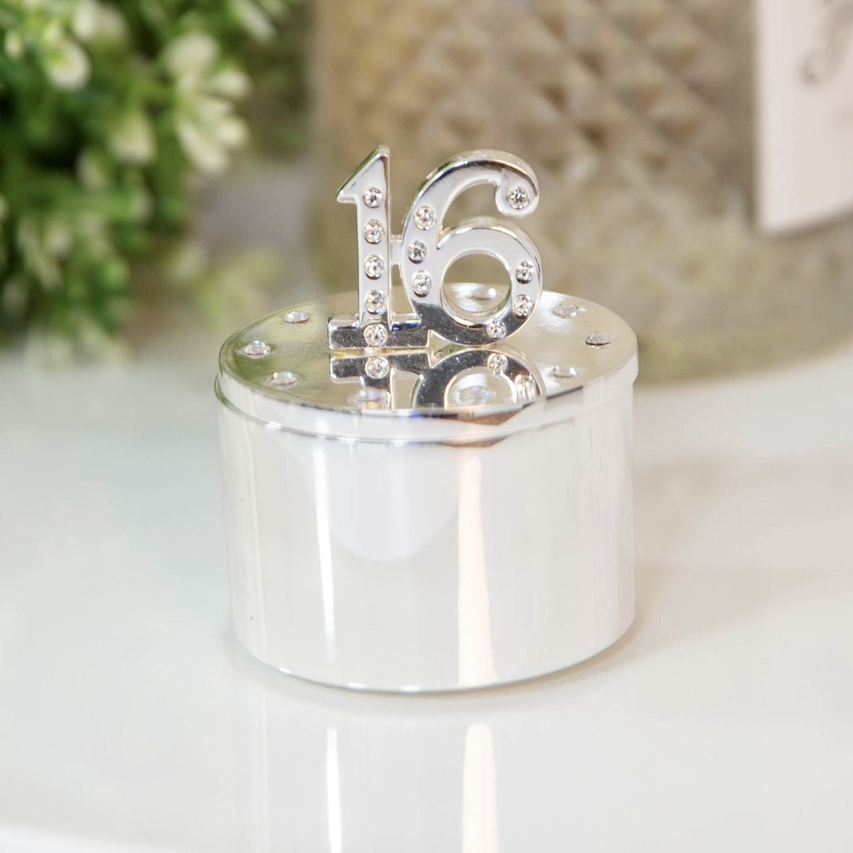 Round silverplated trinket box with a raised, crystal-studded "16" on the lid and crystals set around the edge, polished silver cylindrical base, shown on a light surface.
