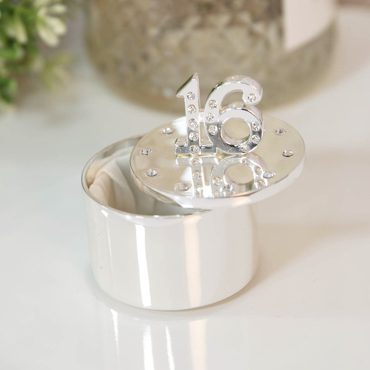 Round silverplated trinket box with a crystal-studded number 16 on the lid, shown open with a cream lining, set on a light surface.