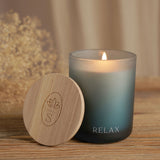 Lit round scented candle in a frosted blue glass, pale at the rim and deeper blue below, with "RELAX" printed in white near the base, next to its wooden lid on a wood surface.
