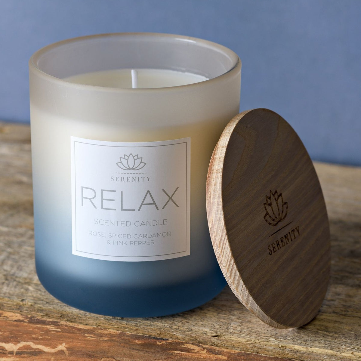 Relax Rose & Spiced Cardamon Scented Candle, 270g, in a round frosted jar shading from white to dusty blue, with a wooden lid resting beside it on a wooden surface.