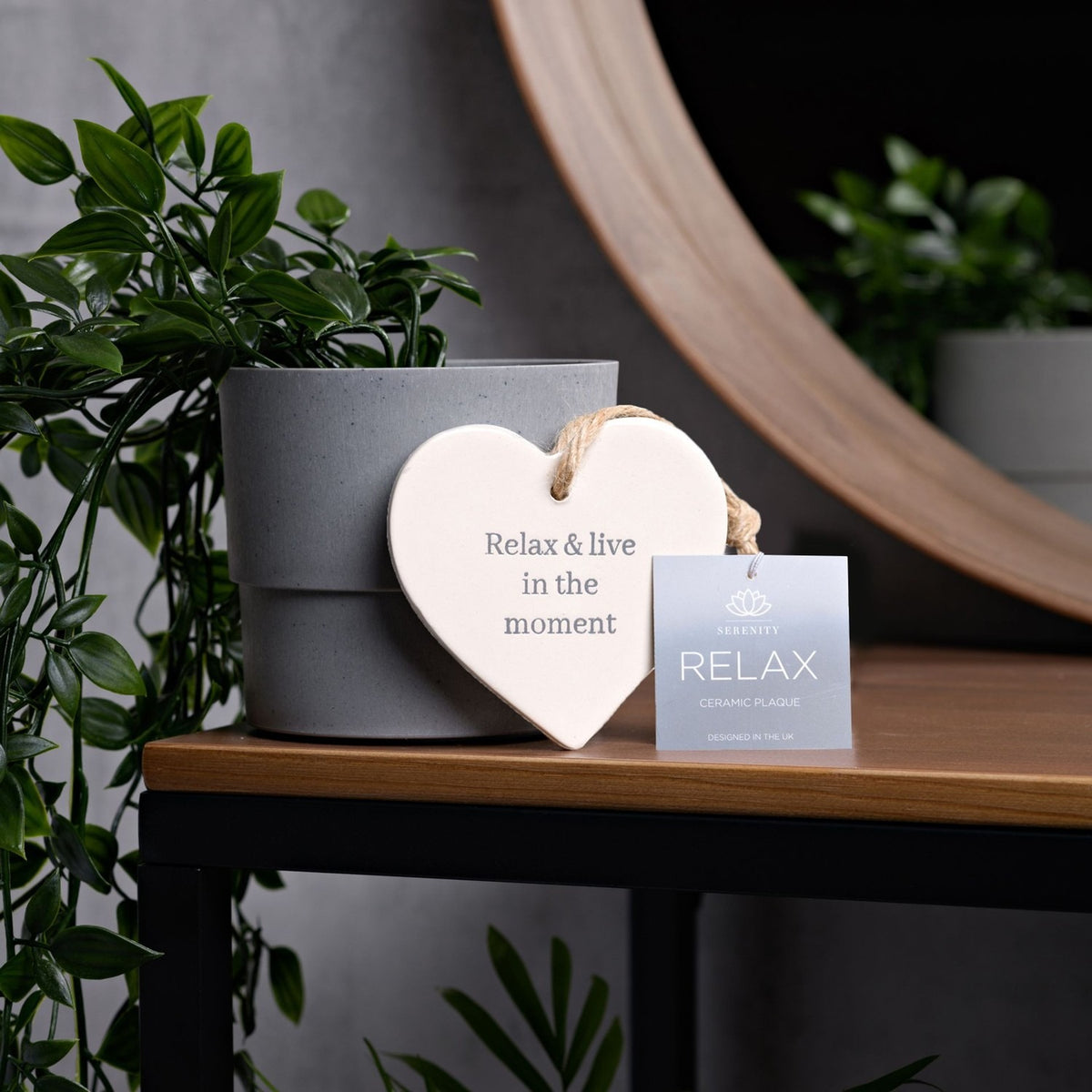 White ceramic heart hanging plaque reading "Relax & live in the moment" with twine loop, leaning against a grey plant pot on a wooden table.