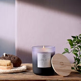 Uplift scented candle in a round purple jar fading from lilac to black, lit wick, with wooden lid beside it on a wooden surface near a pumice stone and plant.