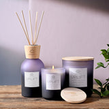 Uplift Dragonfruit Coconut Grapefruit Scented Candle in round purple jars with matching reed diffuser, lit wick visible, with pale wood lids and a lilac background.
