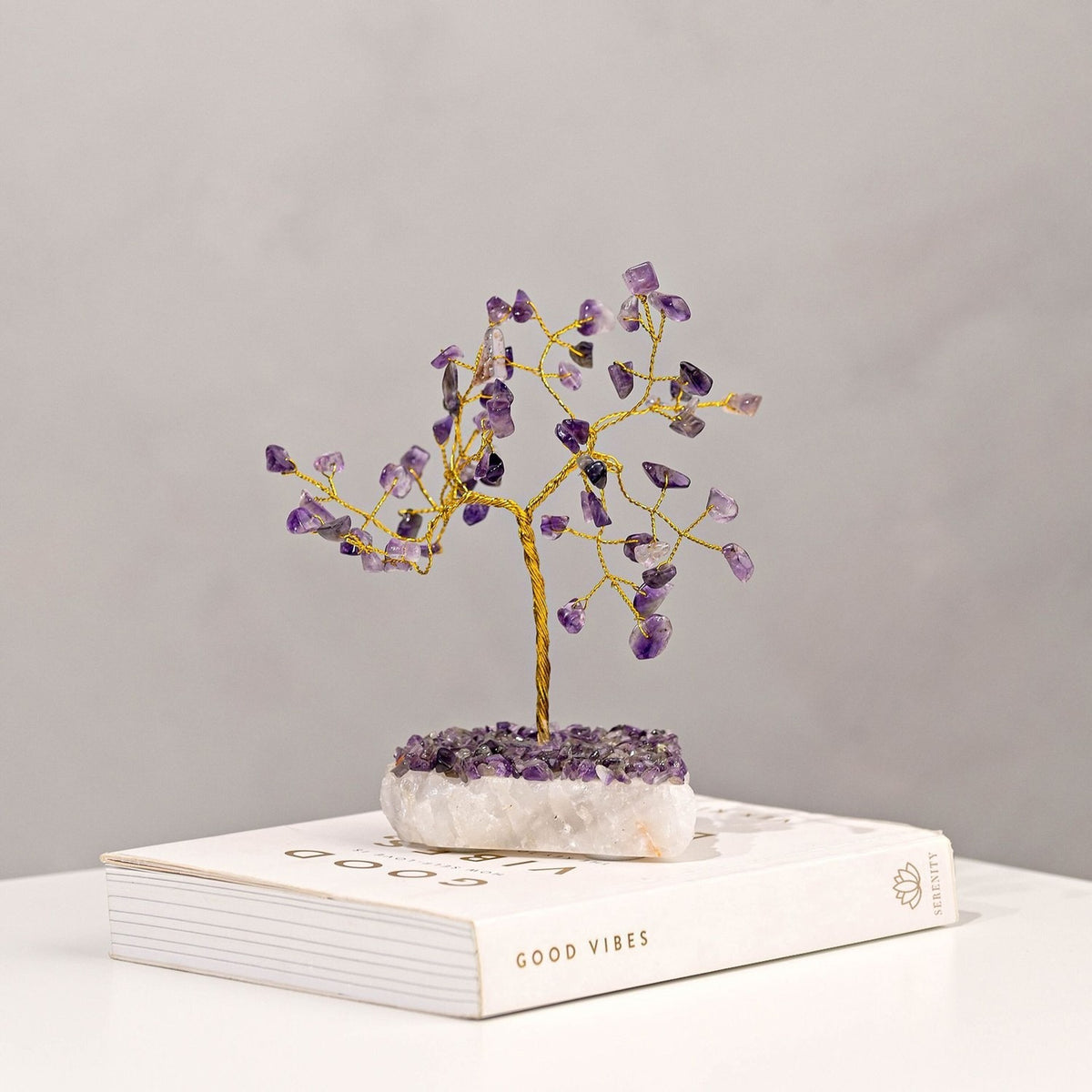 Amethyst Gemstone Tree Ornament with a twisted gold-toned trunk and branches tipped with purple amethyst chips, set on a chunky white crystal base, shown atop a book against a plain grey background.