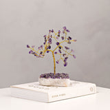 Amethyst Gemstone Tree Ornament with a twisted gold-toned trunk and branches tipped with purple amethyst chips, set on a chunky white crystal base, shown atop a book against a plain grey background.
