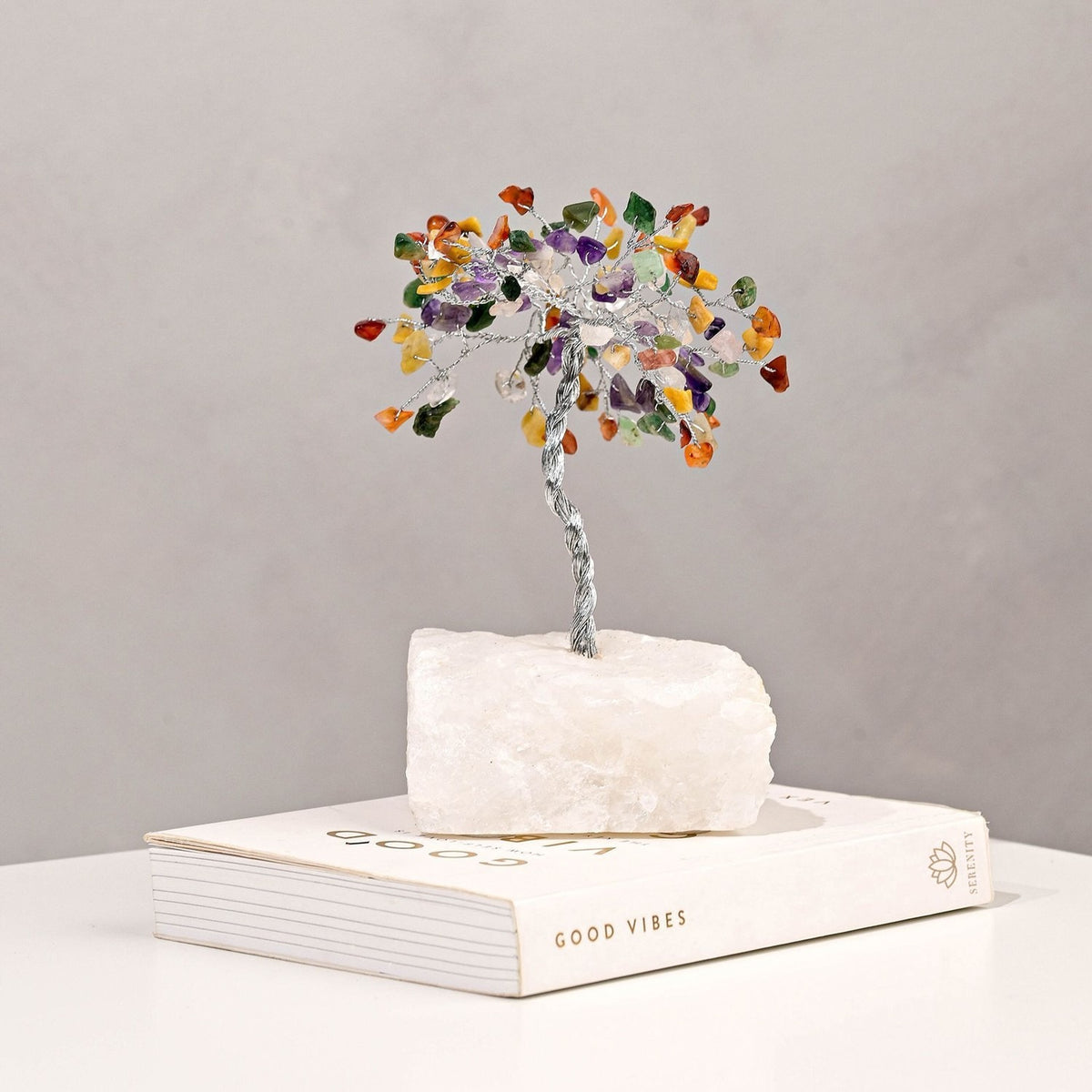 Small multi-coloured gemstone tree ornament with twisted silver-toned trunk and chip canopy on a white crystal base, displayed on a book against a plain grey background.