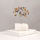 Small multi-coloured gemstone tree ornament with twisted silver-toned trunk and chip canopy on a white crystal base, displayed on a book against a plain grey background.