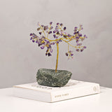 Amethyst Gemstone Tree Ornament with a twisted gold trunk and purple chip canopy on a grey-green stone base, set on a book against a plain light background.