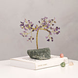 Amethyst Gemstone Tree Ornament with gold twisted trunk and purple chip canopy on a grey-green stone base, displayed on a book against a plain light grey background.