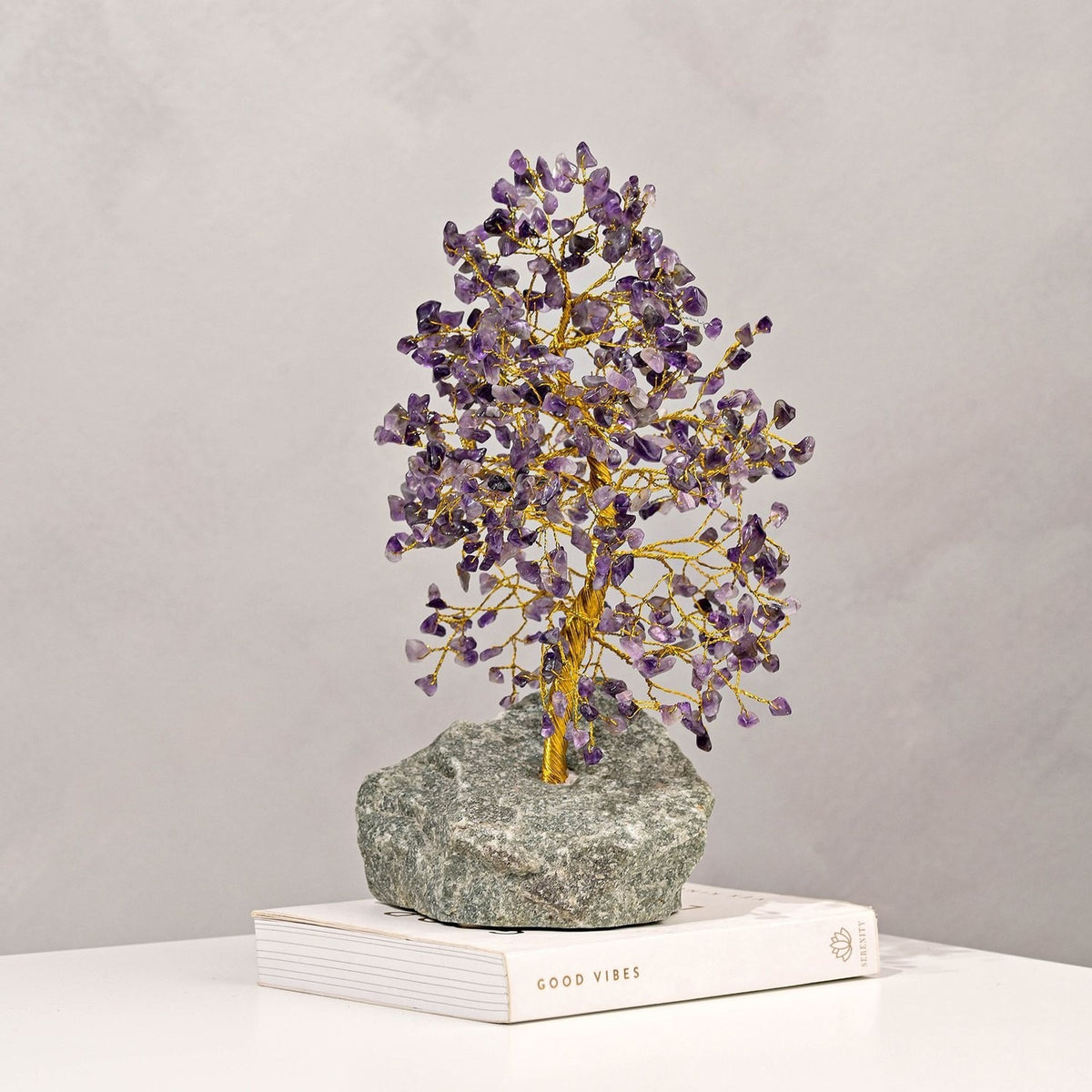 Amethyst gemstone tree ornament with a twisted gold trunk and purple chip canopy, set on a grey-green stone base atop a book, light grey background.