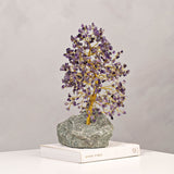 Amethyst gemstone tree ornament with a twisted gold trunk and purple chip canopy, set on a grey-green stone base atop a book, light grey background.