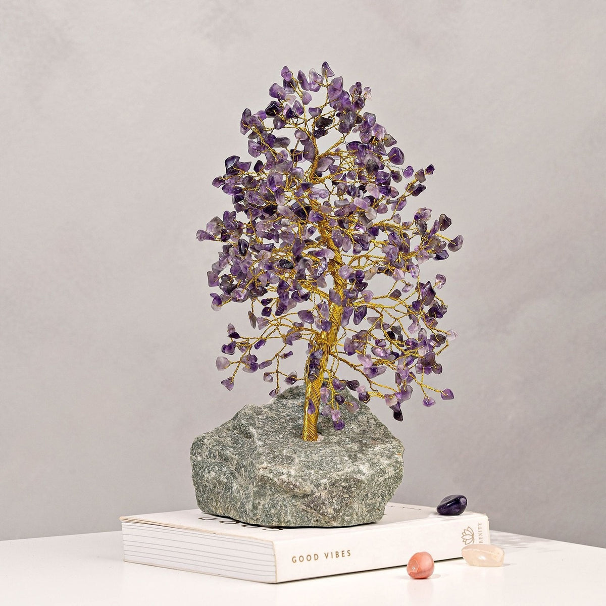 Amethyst Gemstone Tree Ornament with a twisted gold trunk and purple chip canopy on a grey-green stone base, displayed on a book against a plain light grey background.
