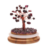 Garnet gemstone tree dome ornament with copper wire branches and purple stone leaves on a bed of chips, set on a round wooden base, shown on a white background.