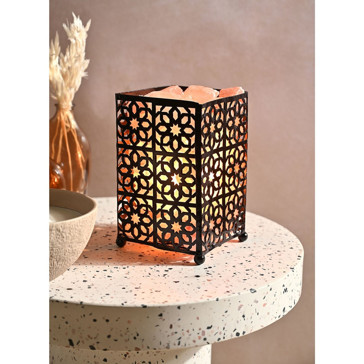 Moroccan style fretwork basket Himalayan salt lamp with black metal rectangular shade featuring star and petal cut-outs, glowing orange, shown on a terrazzo side table.