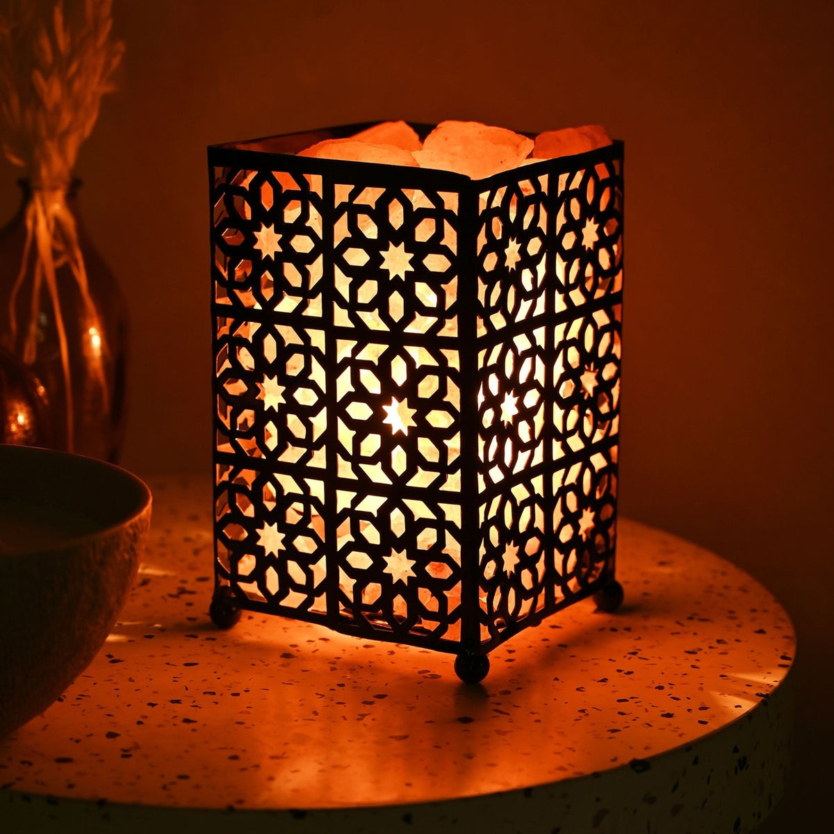 Moroccan style fretwork basket Himalayan salt lamp with black metal geometric star cutwork panels, glowing orange on a speckled side table beside a vase.