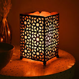 Moroccan style fretwork basket Himalayan salt lamp with black metal geometric star cutwork panels, glowing orange on a speckled side table beside a vase.