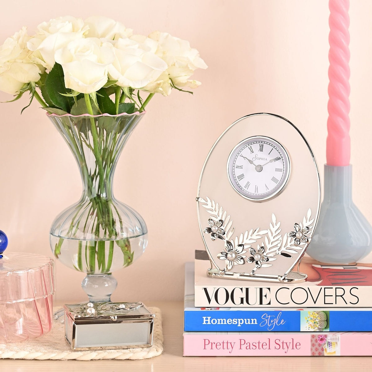 Oval mantel clock with clear glass panel, white Roman numeral dial, and silver leaf and daisy details, displayed on stacked books beside a rose vase.