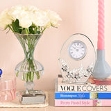 Oval mantel clock with clear glass panel, white Roman numeral dial, and silver leaf and daisy details, displayed on stacked books beside a rose vase.