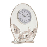 Oval mantel clock with a clear glass panel and silver frame, white Roman numeral dial, and silver leaf sprays with pearl-centred daisies across the base, on a plain white background.