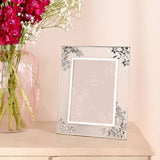 Silver metal rectangular photo frame with daisy clusters and grey blossom detailing at each corner, displayed on a console table beside a vase of flowers.