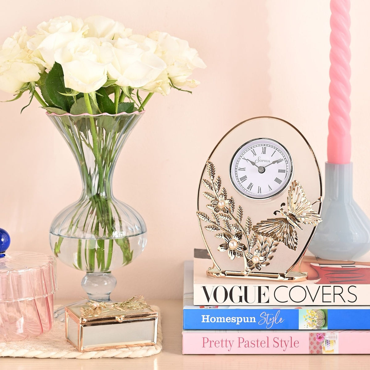 Classic Gold Butterfly and Flower Oval Mantel Clock with clear glass face, white dial and Roman numerals, displayed atop stacked books beside a glass vase of white roses.