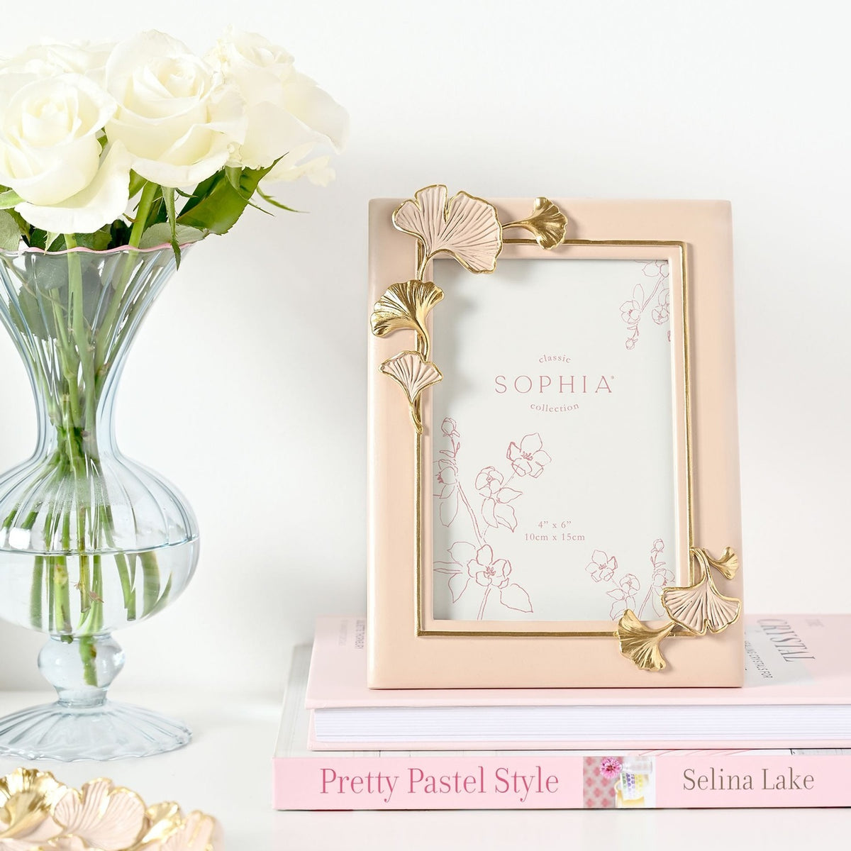 Rectangular resin photo frame in blush pink with gold and pink ginkgo leaf accents at opposing corners, shown on a stack of books beside a vase of white roses.
