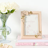 Rectangular resin photo frame in blush pink with gold and pink ginkgo leaf accents at opposing corners, shown on a stack of books beside a vase of white roses.