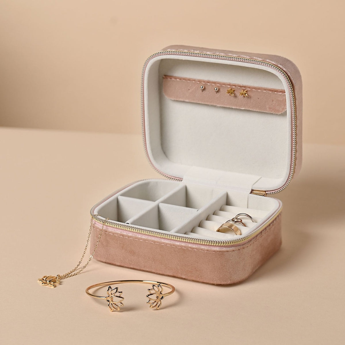 Open pink velvet rectangular jewellery box with gold-tone zip, cream lining, ring slots and compartments, shown with a lotus bangle and pendant necklace.