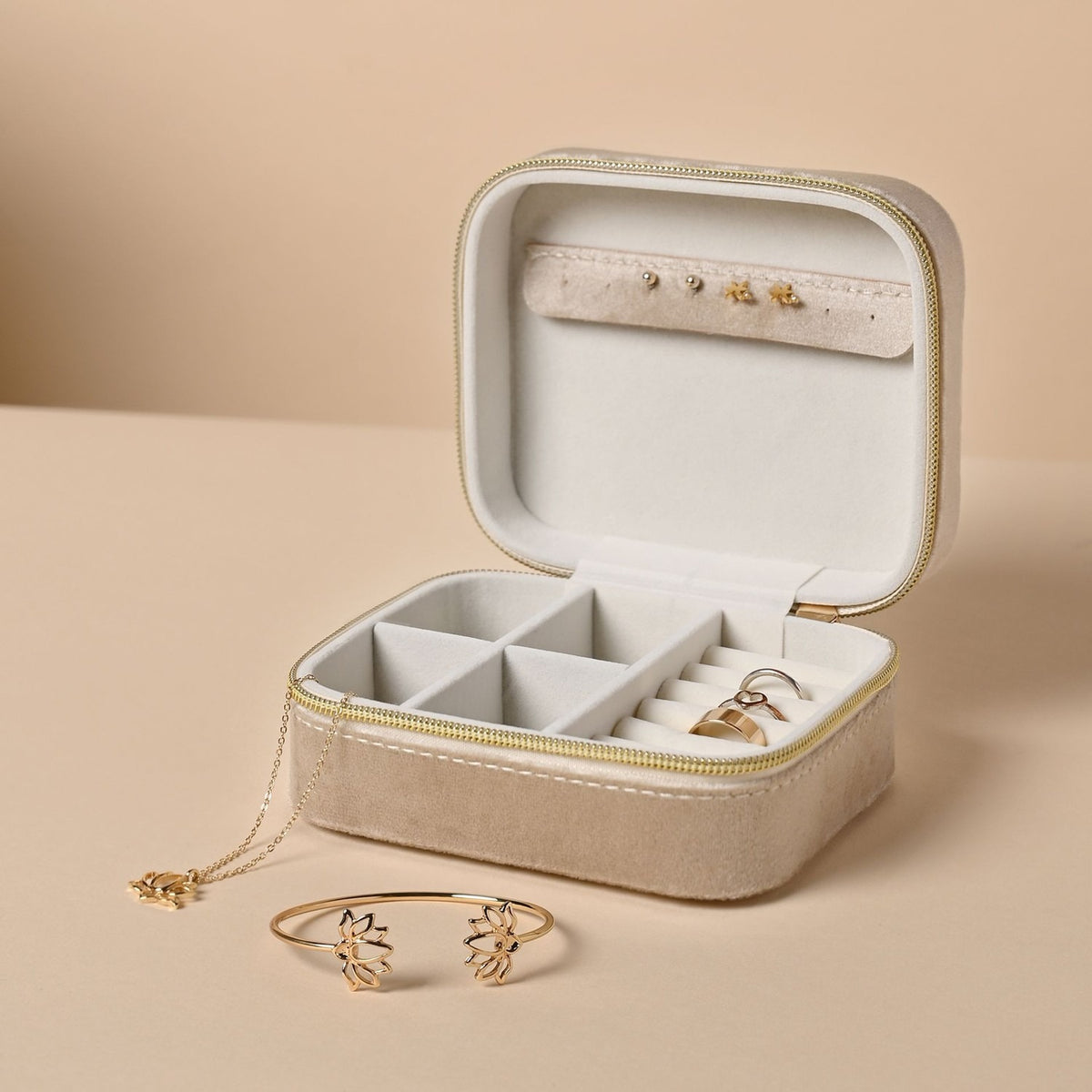 Open rectangular brown velvet jewellery box with gold zip, cream interior, ring rolls, earring strap and compartments, shown with a gold bracelet and necklace on a beige background.
