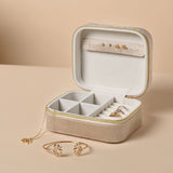 Open rectangular brown velvet jewellery box with gold zip, cream interior, ring rolls, earring strap and compartments, shown with a gold bracelet and necklace on a beige background.