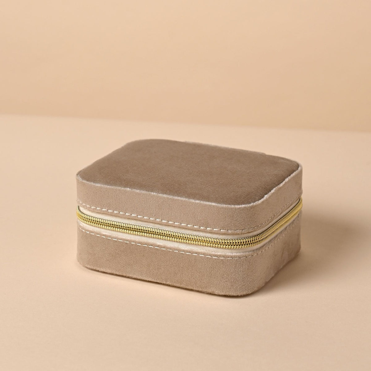 Rectangular brown velvet jewellery box with rounded corners and a gold-coloured zip with pull tab around the front and sides, shown closed on a plain beige background.