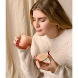 Woman holding the Flow Bergamot, Myrrh and Sandalwood scented candle in a round peach-toned glass, with its wooden lid held in her other hand.
