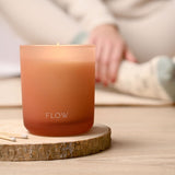 Flow scented candle in a round peach-toned glass with "FLOW" printed near the base, lit on a wood coaster beside matches, shown in a relaxed lifestyle setting.