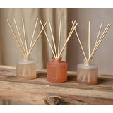 Set of three frosted glass reed diffusers in peach tones labelled Flow, Balance and Breathe, each with natural reed sticks, shown on a wooden shelf.