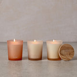 Set of three round frosted glass votive candles labelled FLOW, BALANCE and BREATHE, lit with visible flames, beside a circular wooden lid, on a neutral background.
