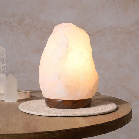Natural white rock salt lamp on a round wooden base, glowing warm amber, shown on a side table beside crystal points.