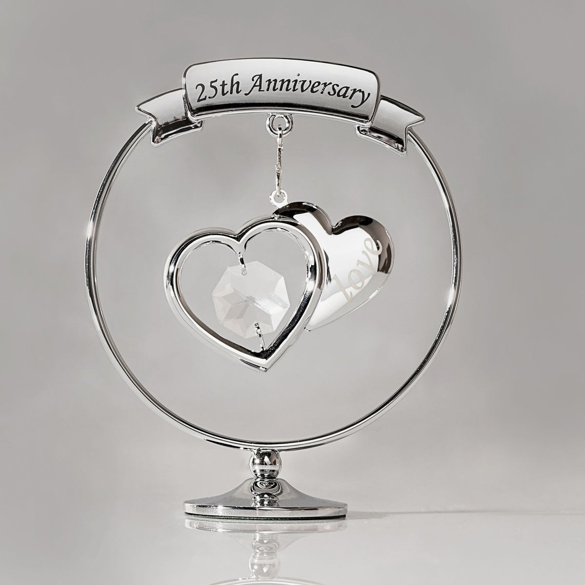 Chrome circle decoration on a round pedestal, featuring a "25th Anniversary" ribbon banner above two overlapping silver hearts, one with a clear crystal drop, set against a plain grey background.