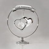 Chrome circle decoration on a round pedestal, featuring a "25th Anniversary" ribbon banner above two overlapping silver hearts, one with a clear crystal drop, set against a plain grey background.