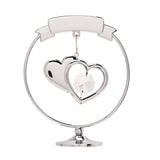 Chrome circle decoration with a curved "25th Anniversary" ribbon banner, two overlapping hearts holding a clear faceted crystal, set on a round silver pedestal base against a plain white background.