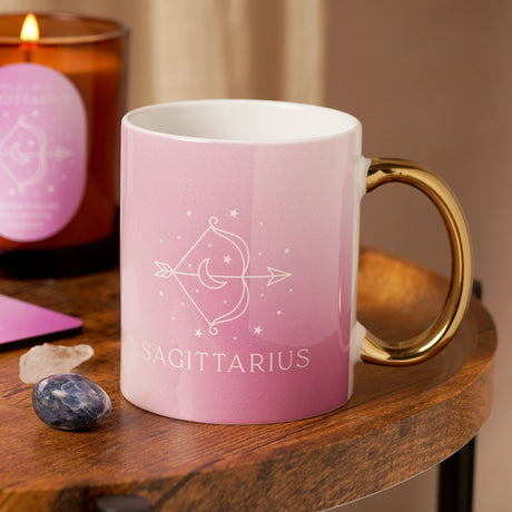 Pink stoneware Sagittarius zodiac mug with glossy finish and gold handle, featuring a white bow-and-arrow design and lettering, shown on a wooden table beside a candle and crystals.