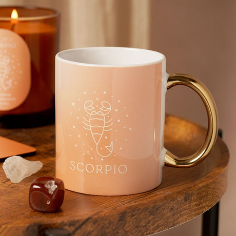 Orange stoneware Scorpio zodiac mug with glossy gold handle and white interior, scorpion line art and star design, shown on a wooden table with candle and crystals.