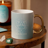 Pale blue stoneware Cancer zodiac mug with a white crab and star motif, "CANCER" text, and a glossy gold handle, styled on a wooden table beside a candle and crystals.