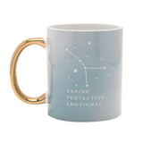 Pale blue stoneware mug with glossy gold oval handle, back view showing white Cancer constellation stars and "CARING PROTECTIVE EMOTIONAL" text, on a plain white background.
