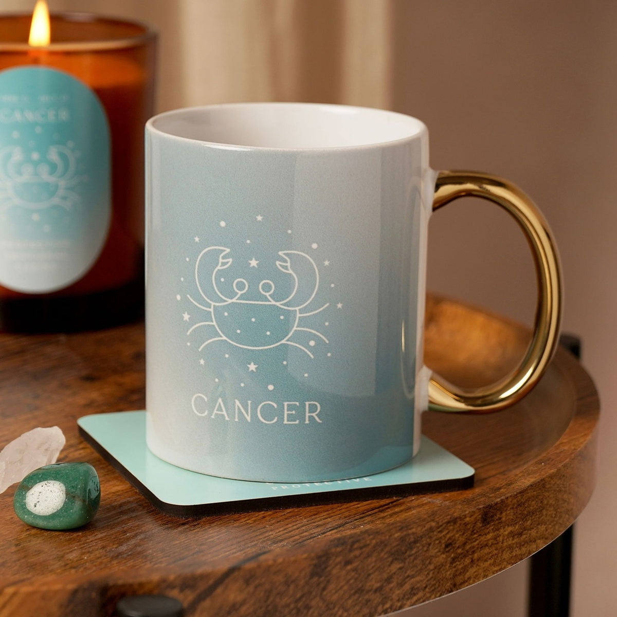 Pale blue stoneware Cancer mug with white crab design and glossy gold handle, shown on a wooden table beside a candle and crystals.