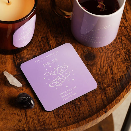 Square glossed wooden Pisces coaster in purple with white twin fish design, dates and traits, shown on a wooden table beside a candle, mug, and crystals.