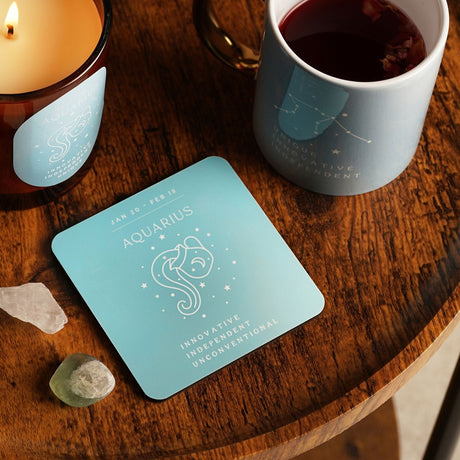 Square wooden Aquarius zodiac coaster in blue with white constellation icon and dates, shown on a wooden table beside a mug, candle, and crystals.