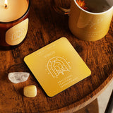 Square wooden Virgo zodiac coaster in golden yellow, with white line art of the maiden under stars, shown on a wooden table beside a candle, mug, and crystals.