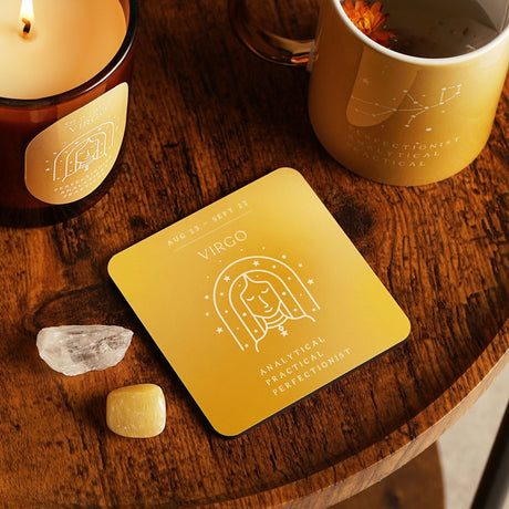 Square wooden Virgo zodiac coaster in golden yellow, with white line art of the maiden under stars, shown on a wooden table beside a candle, mug, and crystals.