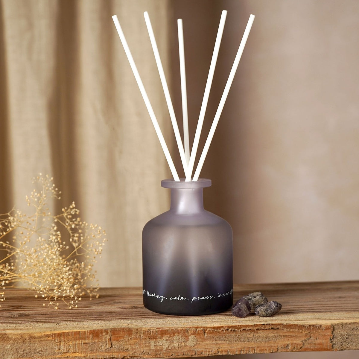Amethyst Crystal Infused Oil Reed Diffuser 230ml in a frosted purple-to-black ombre glass bottle with white reeds and script text, displayed on a wooden shelf beside dried flowers and crystal stones.
