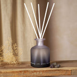 Amethyst Crystal Infused Oil Reed Diffuser 230ml in a frosted purple-to-black ombre glass bottle with white reeds and script text, displayed on a wooden shelf beside dried flowers and crystal stones.